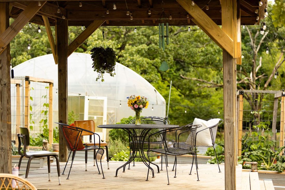 A patio table with a sunflower bouquet under the string-lit pavilion, the high tunnel and garden behind