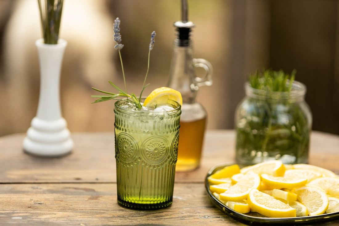 A seasonal mocktail in a green pressed-glass tumbler with lavender, rosemary and lemon beside a plate of lemon slices