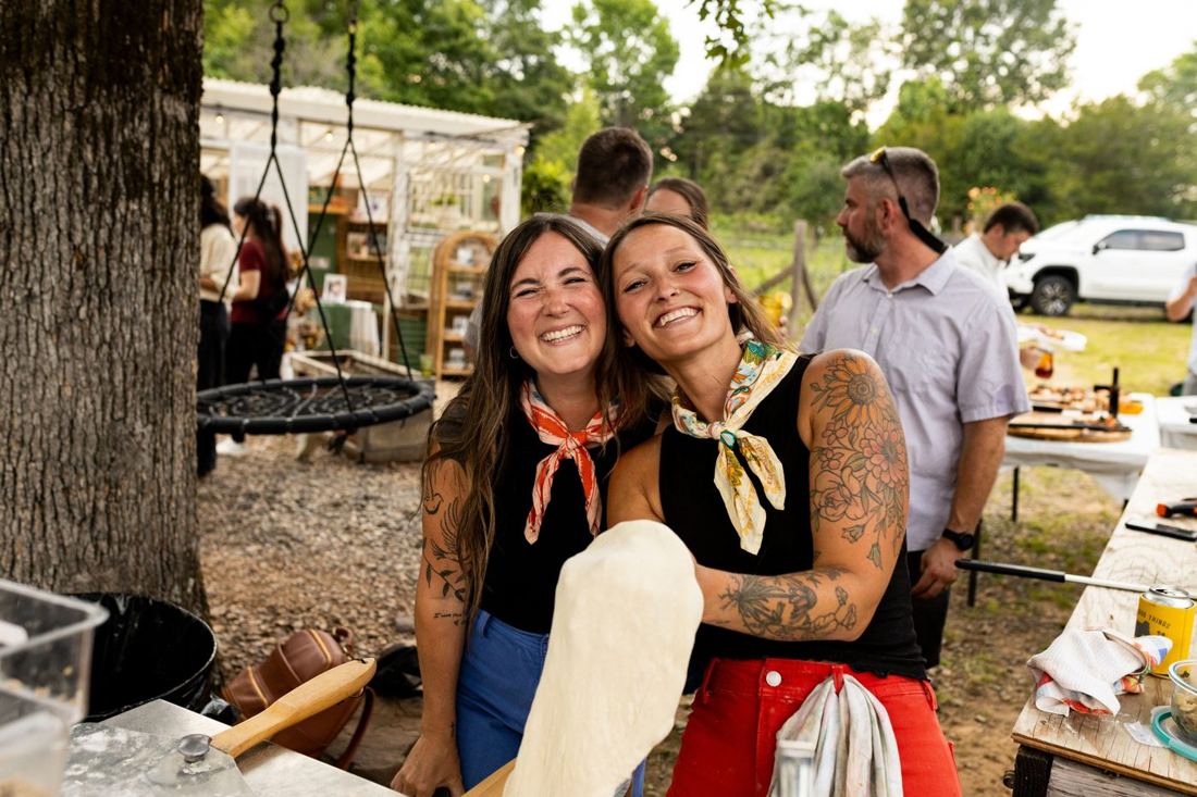 Jill and a friend in matching bandanas grinning over a stretched pizza dough, guests gathered behind them