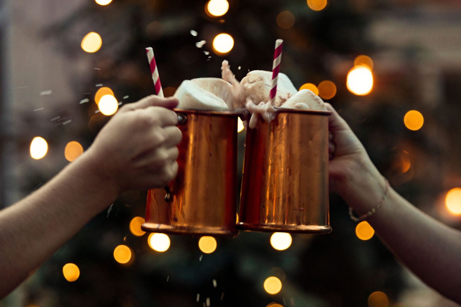 Two hands clinking copper mugs of hot cocoa with marshmallows in front of glowing Christmas lights