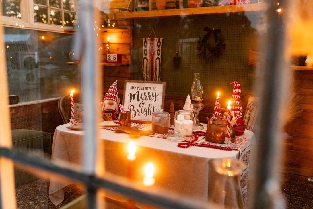Looking through the greenhouse window at the candlelit hot cocoa table with gnomes and a Merry and Bright sign