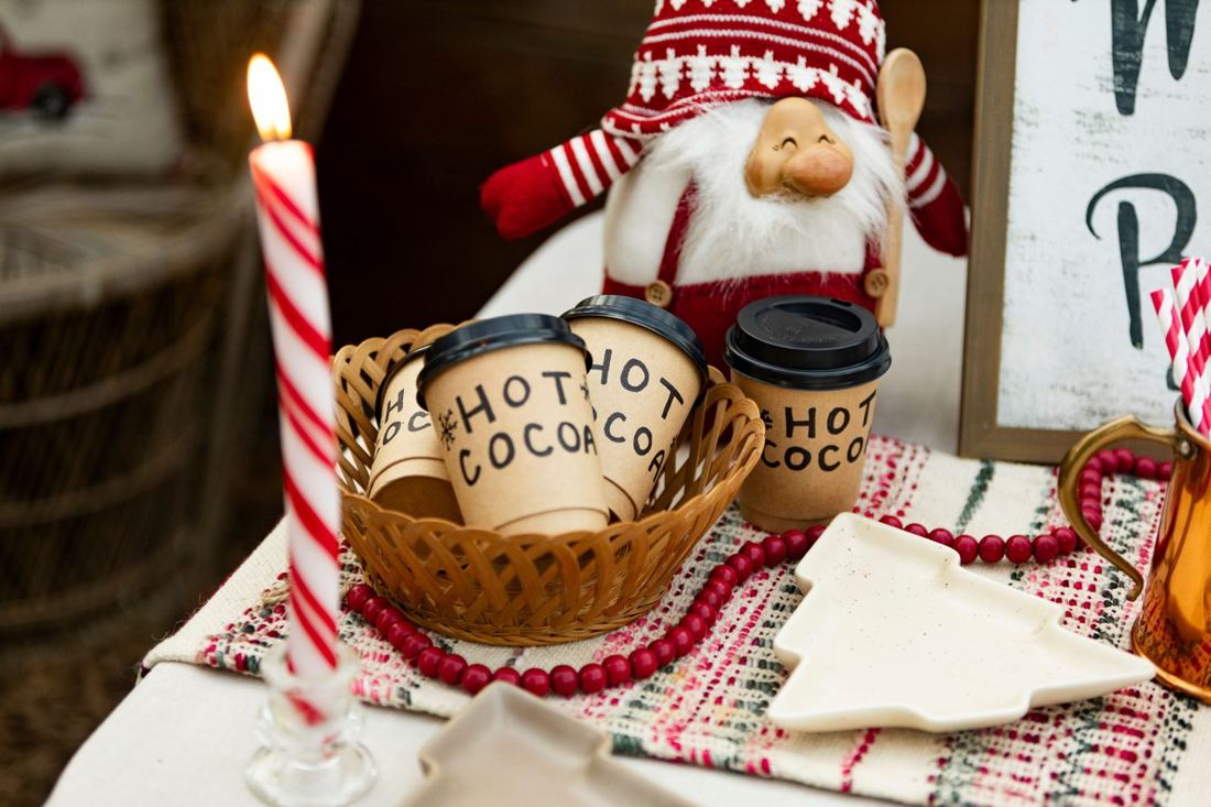 Hand-lettered hot cocoa cups in a basket beside a smiling gnome and a candy-stripe candle