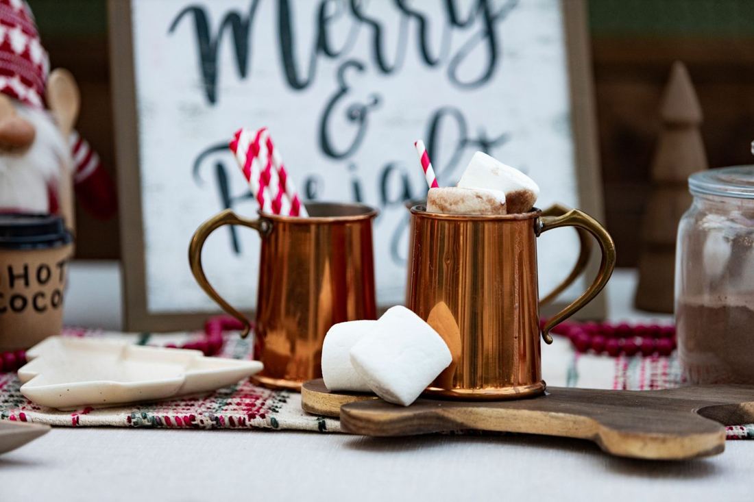 Two copper mugs of hot cocoa with big marshmallows and striped straws in front of a Merry and Bright sign